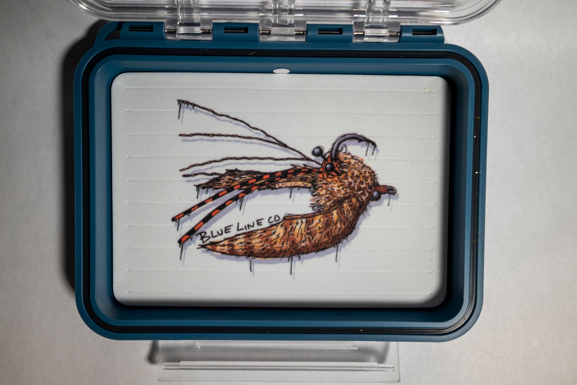 A clear plastic fly box with a blue trim, featuring a crab pattern illustration on the lid.