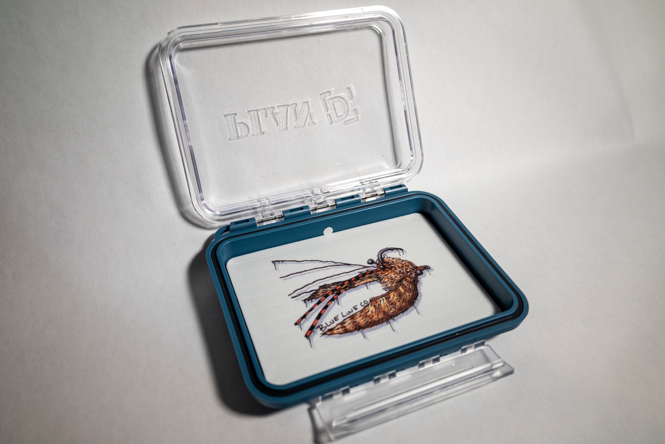 A clear plastic fly box with a blue trim, featuring a crab pattern illustration on the lid.