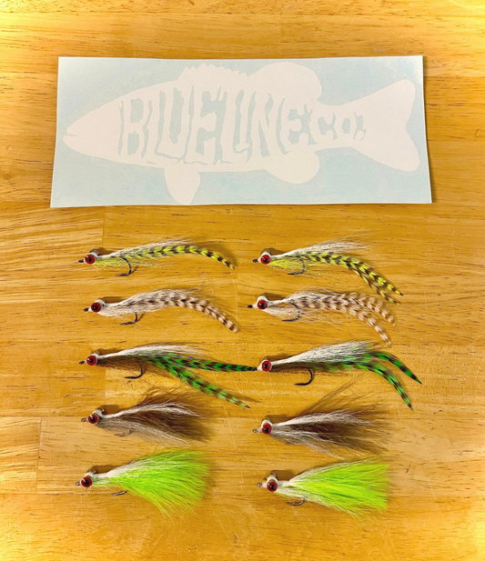 A collection of hand-tied fishing lures resembling clouser minnows, displayed on a wooden surface. The lures come in various colors including green, gray, and white, and are arranged in rows with the BluneLineCo brand sticker at the top.